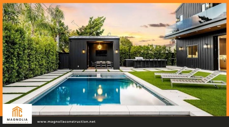 Modern backyard featuring a rectangular swimming pool, a black outdoor cabana with a dining set, lounge chairs, and a two-story dark vertical siding ADU, branded by Magnolia Construction.